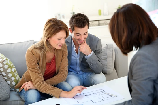 Couple assis sur canapé gris consultant des documents avec une conseillère professionnelle dans un salon moderne lumineux