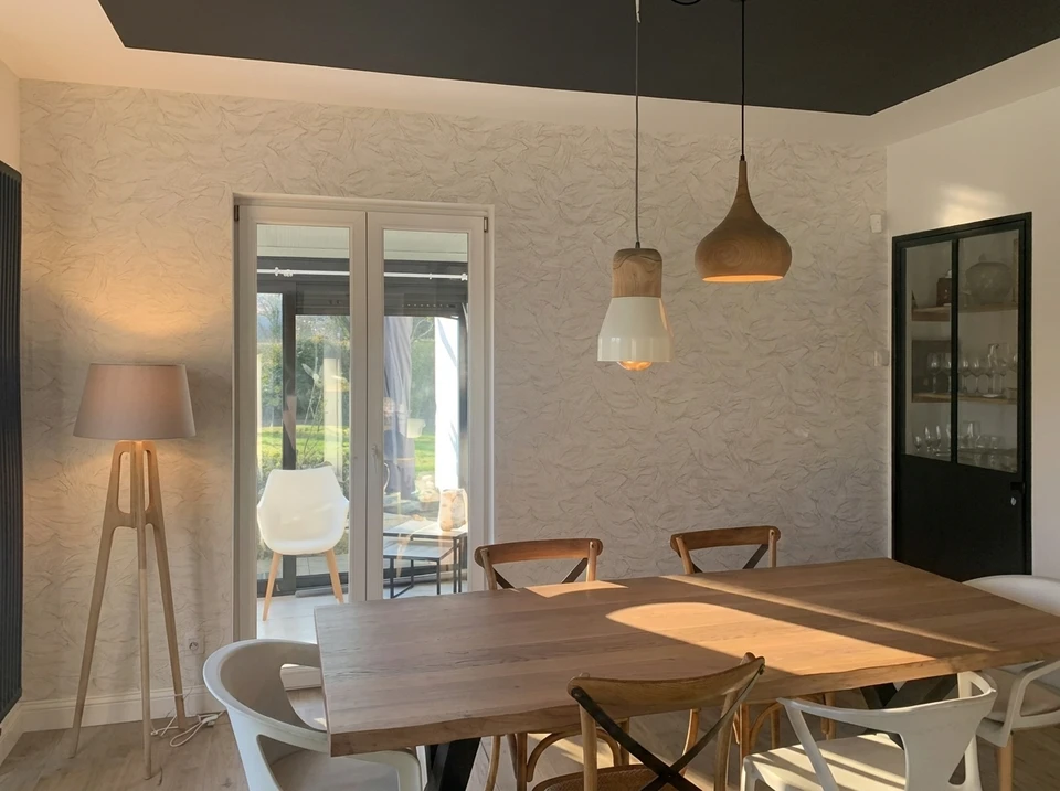 Artisan en salopette et t-shirt blanc installe du papier peint sur mur intérieur résidentiel, appliquant la bande avec les mains près de l'angle et plinthes