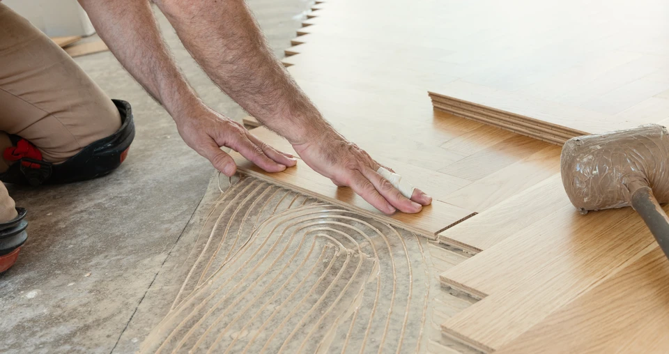 Artisan à genoux installant du parquet dans une pièce blanche, mesurant une lame avec un mètre ruban
